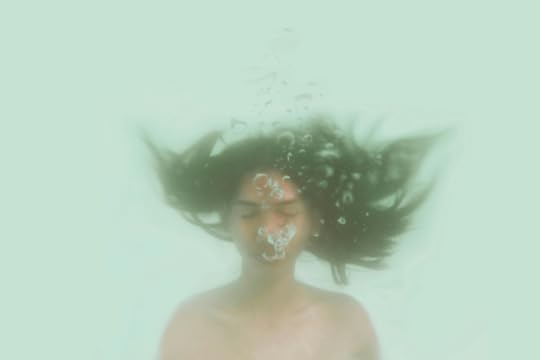 Image: a woman is underwater with her eyes closed, exhaling and releasing air bubbles that float upward.