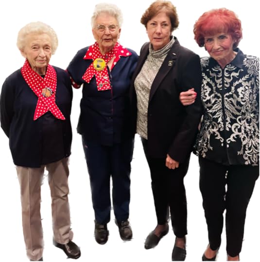 Four women standing side by side against a black background. From left to right: the first woman has white hair, wears a navy blue blazer over a beige pair of pants, and has a red scarf with white polka dots tied at her neck. The second woman has light gray hair, wears a navy blue blazer with red details and a matching red scarf with white polka dots, and sports a button on her lapel. The third woman has brown hair, is wearing a black coat over a patterned shirt, and her posture suggests she may be in mid-motion. The fourth woman has red hair, wears a black and white patterned blazer over black pants, and has her arm linked with the third woman. 