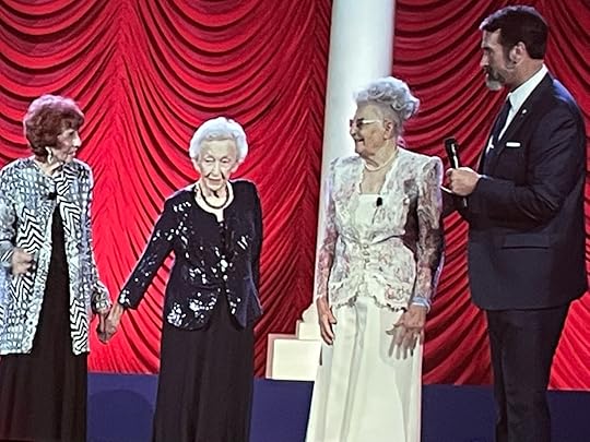  four individuals on the stage; from left to right, there are three women and a man holding a microphone. The first woman is wearing a black and white chevron patterned jacket, the second is in a black dress with sparkling embellishments, and the third is dressed in a light-colored outfit with lace details. The man on the far right is dressed in a dark suit and tie. All individuals are facing towards an unseen audience, and there appears to be a column partially obscuring the view of the third woman.