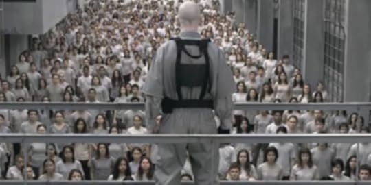 A still from the first season of 3 Percent on Netflix, showing a bald man wearing a harness, standing on a walkway looking down at a sea of young people wearing identical white clothes