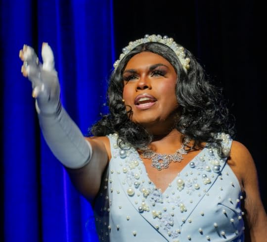 A drag queen sits on a stool with arms outreached