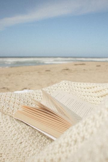 open book lying on top of a cream colored blanket spread on the sandy beach with the ocean in the background.