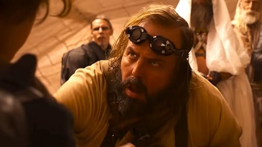 The Organic Mechanic with goggles on his forehead looks at someone inside a tent in Furiosa
