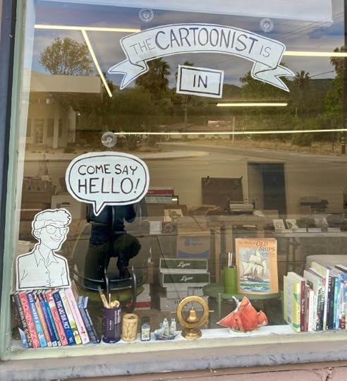 A cardboard cutout of Lucy saying "come say hello" in a window under a similar black and white sight reading "the cartoonist is in."