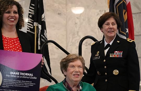 four individuals on the stage; from left to right, there are three women and a man holding a microphone. The first woman is wearing a black and white chevron patterned jacket, the second is in a black dress with sparkling embellishments, and the third is dressed in a light-colored outfit with lace details. The man on the far right is dressed in a dark suit and tie. All individuals are facing towards an unseen audience, and there appears to be a column partially obscuring the view of the third woman.