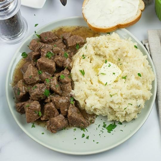 A plate with beef tips and gravy served with mashed potatoes on a dinner plate. Sliced bread in the background.