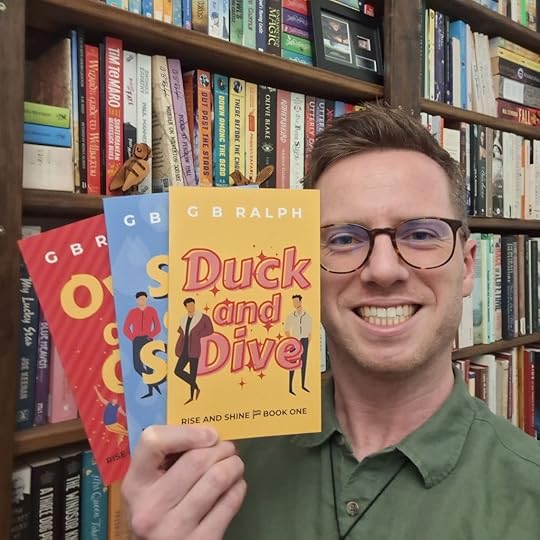 Author holding up books in front of bookshelves. First book is Duck and Dive by G B Ralph.