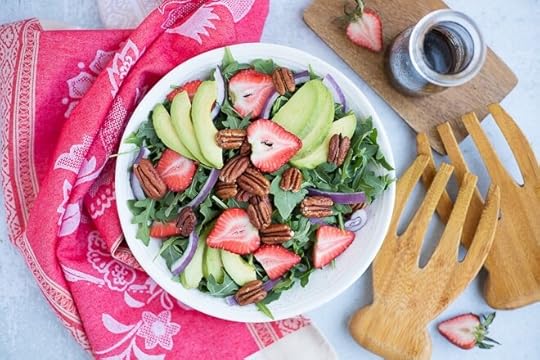 white bowl with strawberry salad and jar of balsamic dressing next to wood serving spoons and red linen