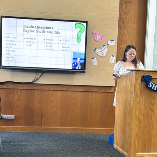 Penny stands at a classroom podium next to a TV with her presentation.