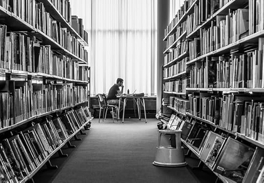 View down a set of library shelves to a man reading at a desk in front of a window.