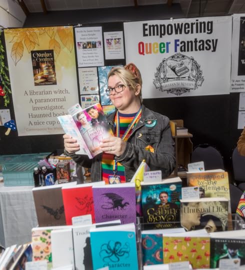 A blonde person wearing tiger ears and a rainbow shirt smiles confidently. They are seated among an array of fantasy books,
