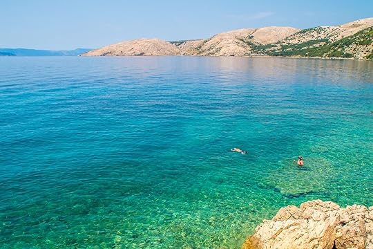 Crystal clear blue green Adriatic sea and a rocky beach on the island Krk, Croatia, with some tourists snorkeling and swimming, with other islands in the background