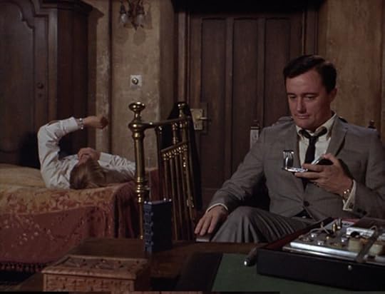 Napoleon sits at a small desk while Illya lies on the only bed in the room, looking at his watch