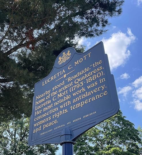 Blue historical marker indicating nearby location of Lucretia Mott's home, Roadside, with blue sky and leafy tree branches in background.
