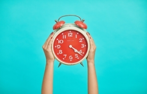 Two hands holding up an orange alarm clock while demonstrating that writing habits take time to build!