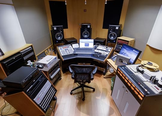 Wide-angle view of a recording studio. One swivel chair sits in front of an audio control board with speakers and other instruments surrounding it.