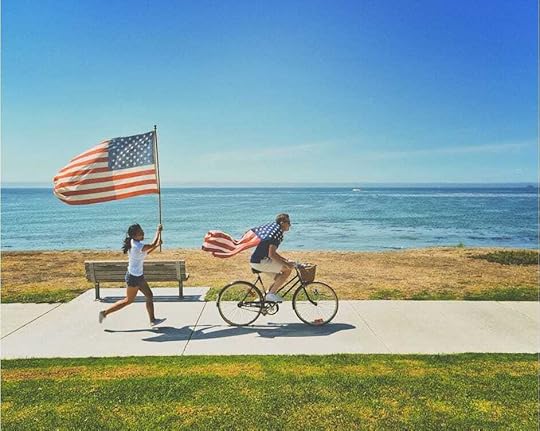 Woman holding American flag high while chasing bike ridden by a man wearing the American flag over his shoulders.