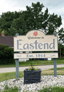 Wooden sign at the entrance to Eastend, Saskatchewan has vintage gray lettering on cream background, decorated with red tiger lily, reading Welcome to Eastend, Est. 1914.