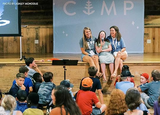 Penny sits on stage between two young adults. She holds a mic and is singing while a group of kids sits in front of her listening.