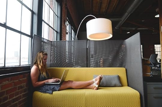 Woman with long blonde hair stretched out on a mustard colored couch working on a laptop. Her back is to a window in an industrail space, and she's partitioned off with a large mesh screen.