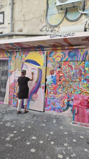 The artist paints the eye of a girl drawn on a wall. the girl is smmiling , wears a yellow hat with a blue band. on the nearby wall colorful flowers