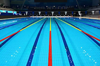 Olympic swimming pool, filled with water but no people. Blue stripes on bottom of pool. Lane dividers red, then yellow