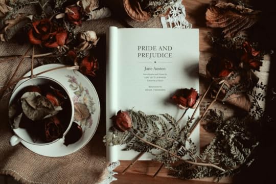 Image: a copy of Pride and Prejudice by Jane Austen open to the title page on a table surrounded by dried Autumn leaves and roses and a teacup.