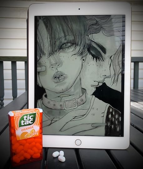 The cover of A+E, which doesn't even have the author's name or title on it, on an iPad. It depicts two gothy, gender-ambiguous teenagers in an embrace. In front of it, sitting on a picnic table, is a package of orange tic tacs, some of them on the table itself. I had to chase a cat off the picnic table to get this shot, and he used this opportunity to sneak into my house and start eating my cat's food, so I hope you appreciate my sacrifice here.