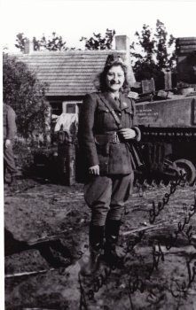 A black and white photograph of a woman standing outdoors in front of a house. She is wearing a military uniform with a belt and boots, and she is smiling. The background includes a vehicle, trees, and part of the house. The image has a vintage or historical appearance, suggesting it may have been taken during a past era. There is some handwritten text on the lower right side of the photograph