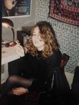 Picture of Kathryn Mockler, a white teenager with brown curly hair sitting at a white desk with her eyes closed holding a cigarette. She wears all black. A white and black patterned scarf hangs on the wall beside a Pink Floyd poster.