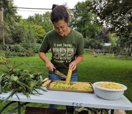 woman standing at a table cutting corn off the cob with a bowl full of kernels of corn.