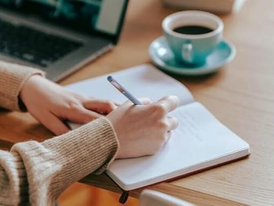 Person writing in a notebook with a laptop nearby, focused on creating an engaging hook for a novella.