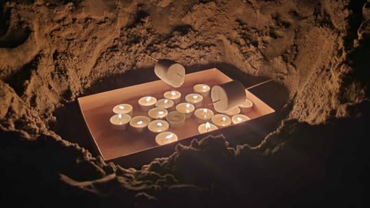 A collection of lit tea candles in a box, set into a pit in the sand, with two huge marshmallows on bamboo sticks stuck into the sides of the pit.