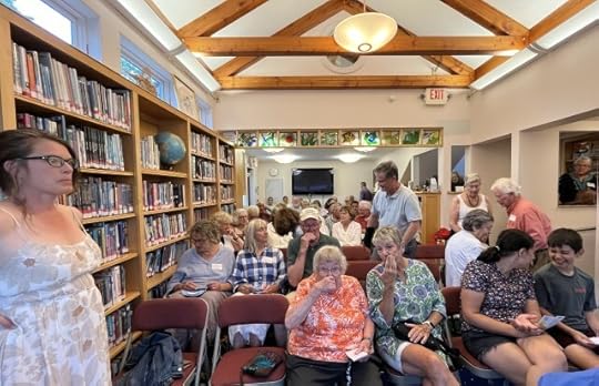 A library room, with a wall of books on one side and packed with people sitting in folding chairs