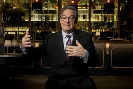 A man in a suit and tie gestures as he speaks, sitting in front of a bar