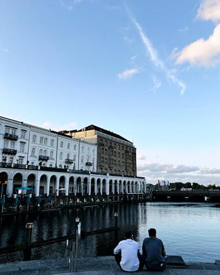Die Hamburger Alsterarkaden und die Kleine Alster. Zwei Menschen sitzen auf den Treppen am Ufer, man sieht sie von hinten. Sommerliche Anmutung, blauer Himmel, Dekowölkchen.