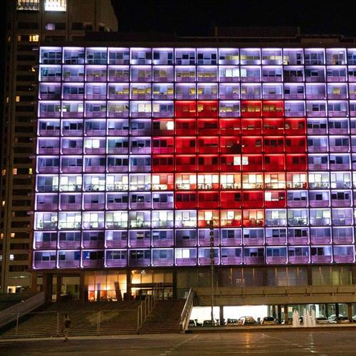 A building at night with its windows illuminated to form a large red heart on a background of purple and white lights. The structure is tall and rectangular, with multiple floors, creating a striking visual display in the dark. The lights create a vibrant, colorful pattern that stands out against the night sky, symbolizing love.