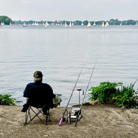 Blick auf die Außenalster von der St-Georg-Seite aus. im Vordergrund ein Angler am Ufer, er sitzt in einem Klappstuhl, bene sich zwei aufgestellte Angeln. Im Hintergrund etliche weiße Segel.