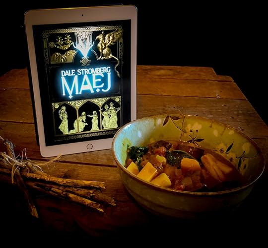 An ebook copy of Maej by Dale Stromberg on an iPad. It's sitting on a rustic wood coffee table. There's a bowl of vegetable stew and some sticks bundled together.
