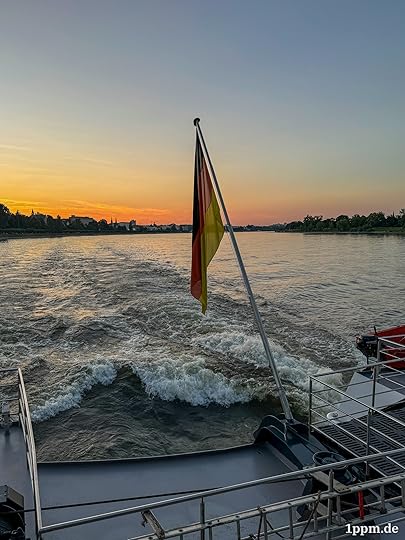 Von hinten von einem Schiff fotografiert, man sieht die Wellen, die das Schiff aufwirft. Außerdem eine Deutschlandflagge und im Horizont einen gelb-orangenen Sonnenuntergang.
