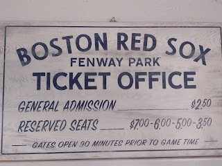 Wood sign that says in blue lettering (against faded white backdrop): Boston Red Sox Fenway Park Ticket Office. General Admission $2.50. Reserved seating $7.00 - $6.00 - $5.00 - $3.50. Gates open 90 minutes prior to game time