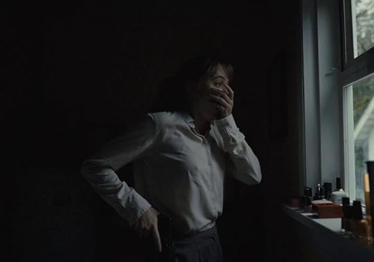 Maika Monroe holds her left hand to her mouth, in a gesture of fear and disgust, and draws her gun with her right while looking out a window in