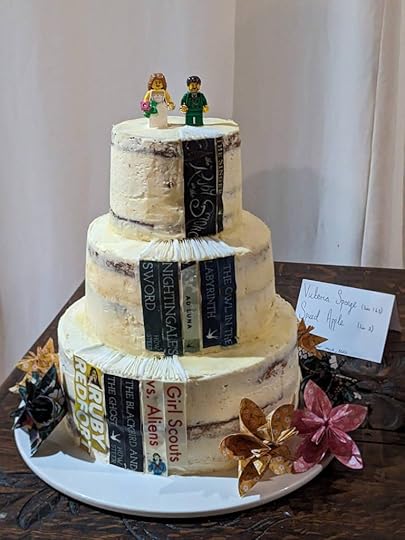 A 3-tiered wedding cake, topped with LEGO minifigures of the bride and groom. Decorating the sides are the spines of books.