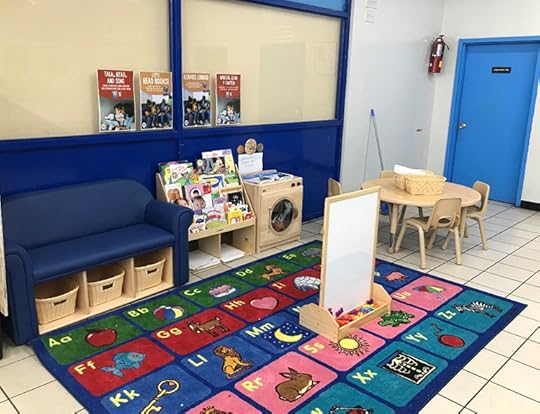 Color photo of a pre-school type reading space, with a colorful mat of the alphabet, a whiteboard, a small round table with charir and books in a rack.