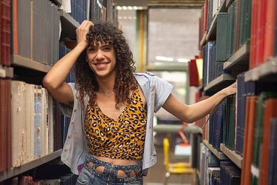 Camila, a Brazilian trans teenager played by trans actor/performer Nila, poses in a cute leopard print shirt and sweater in the middle of library bookshelves in season three of Back to 15