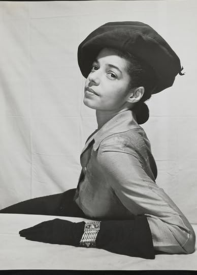 Half length shot of a woman seated sideways to camera, posing in front of a dropcloth and wearing a stylish hat and gloves.