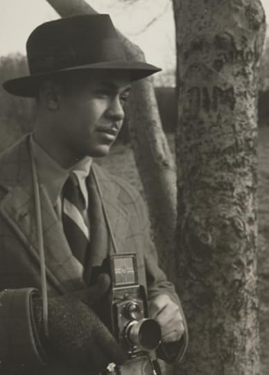 Ralph Ellison, wearing a suit, tie and fedora, with a camera in St. Nicholas Park, New York City.