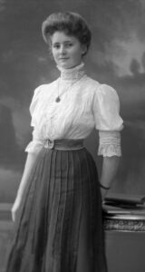 Smiling young woman in black and white photo wears high-necked lace blouse with locket around her neck and dark pleated skirt, her hair in a bouffant chignon