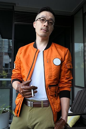 Photo of me dressed as Kim Kitsuragi from Disco Elysium. I'm a middle-aged Asian guy in an orange bomber jacket, olive pants, round glasses, and brown leather driving gloves. Waist-up shot. I'm standing with my head tilted to the side, with the cigarettle held at my waist.
