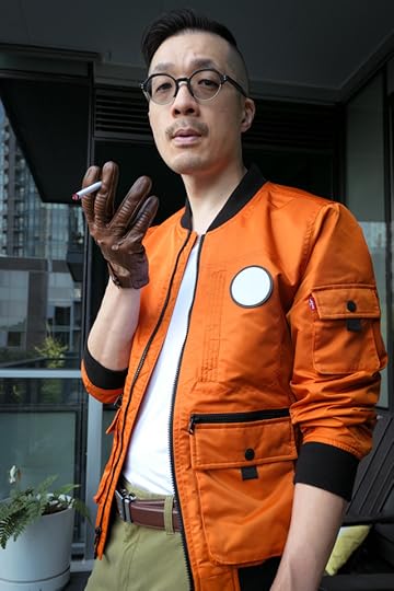 Photo of me dressed as Kim Kitsuragi from Disco Elysium. I'm a middle-aged Asian guy in an orange bomber jacket, olive pants, round glasses, and brown leather driving gloves. Waist-up shot. I'm facing sideways, but with my head turned towards the camera. I'm cradling the cigarettle with my palm up.